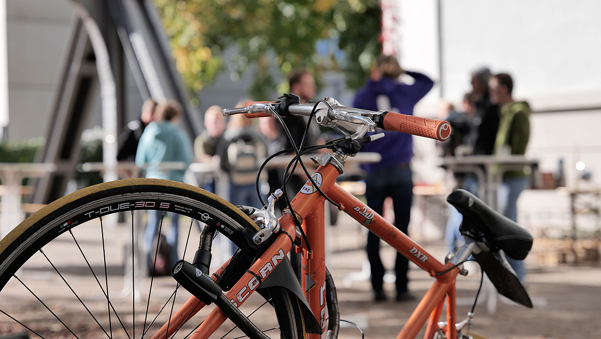Kaputtes Fahrrad, im Hintergrund Studierende an Stehtischen beim Kaffeetrinken